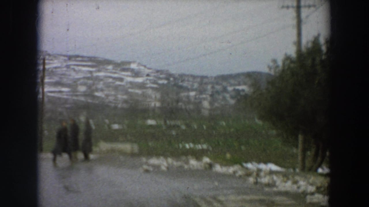 1958 Italy home-movie frame showing three people in dark coats walking on a damp rural road with terraced snowy hills behind them.