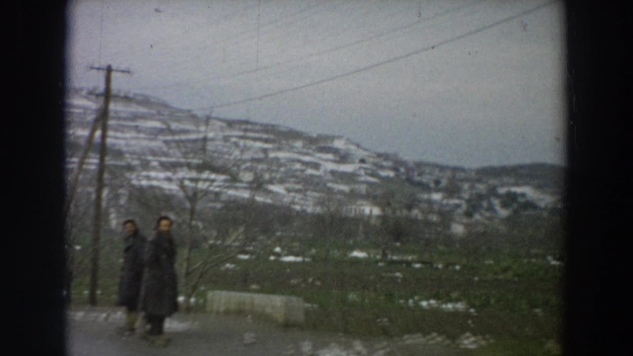 Two dark-coated pedestrians appear beside a rural Italian road, with utility poles, bare trees, and snow-dusted terraced hills in the background.