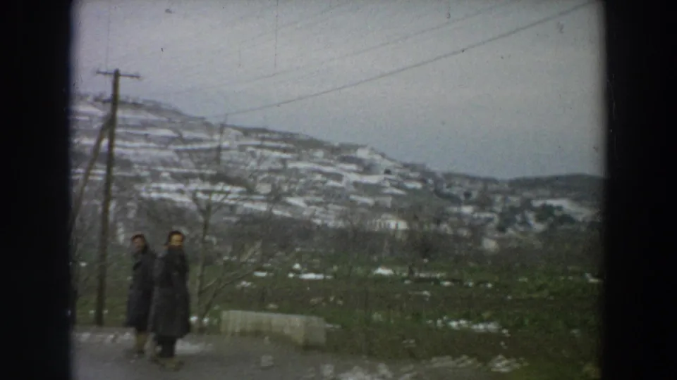 Two dark-coated pedestrians appear beside a rural Italian road, with utility poles, bare trees, and snow-dusted terraced hills in the background.