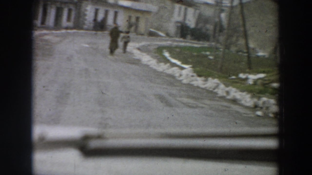 The moving 8mm view looks past part of a car toward a wet village road, distant pedestrians, low buildings, poles, and patches of snow.