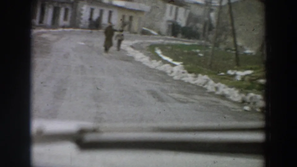 The moving 8mm view looks past part of a car toward a wet village road, distant pedestrians, low buildings, poles, and patches of snow.