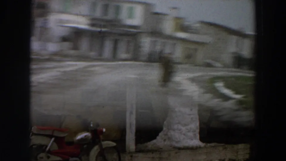 1958 Italy 8mm frame with a blurred view past dark edges toward a snowman, yellow stucco building and red scooter.