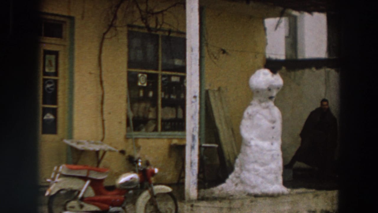 1958 Italy home-movie frame showing a lopsided snowman outside a yellow stucco house, with a red scooter and distant person.