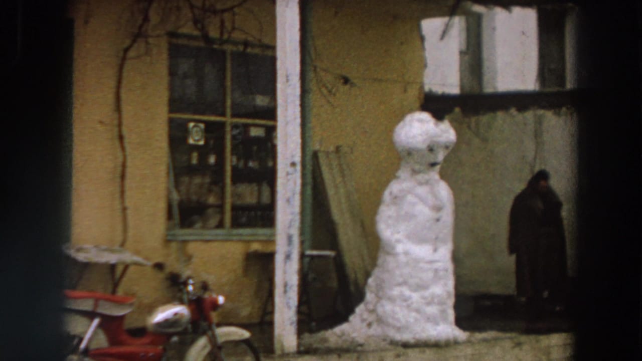 1958 Italy 8mm frame of a handmade snowman by a yellow house, red scooter at left and a bundled person in the background.