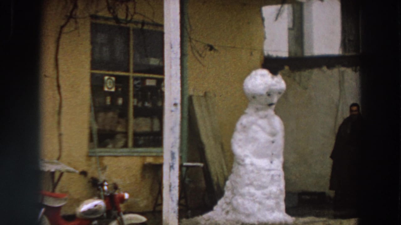 Final 1958 Italy frame holds on the snowman in front of a yellow stucco exterior, with scooter details and a distant person at right.