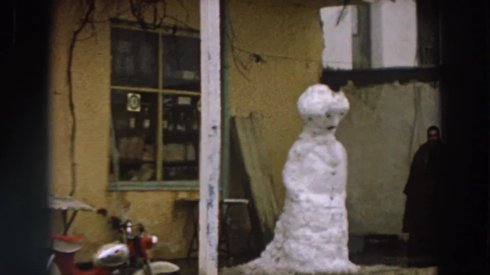 Final 1958 Italy frame holds on the snowman in front of a yellow stucco exterior, with scooter details and a distant person at right.