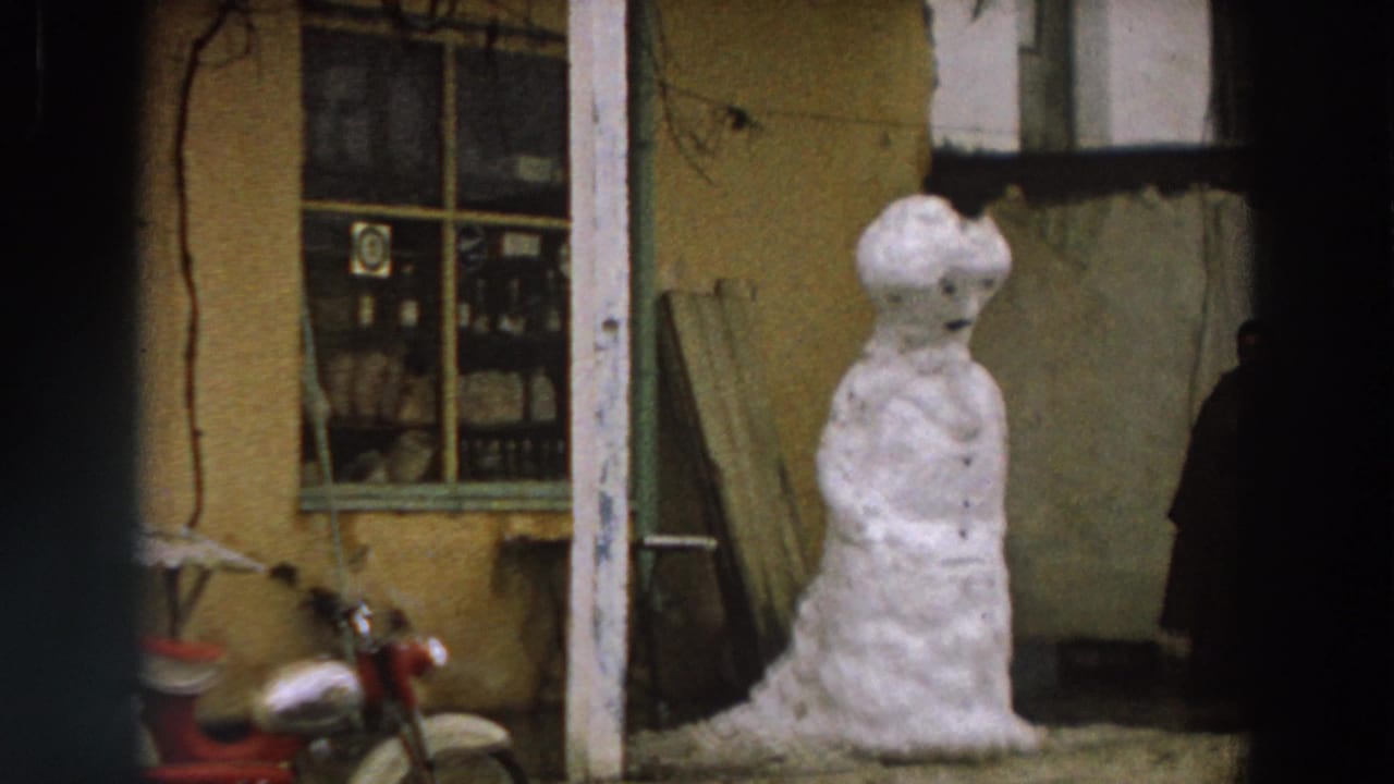 1958 Italy home-movie frame showing a snowman beside a weathered building, shop-style window, and partly visible red motorcycle.