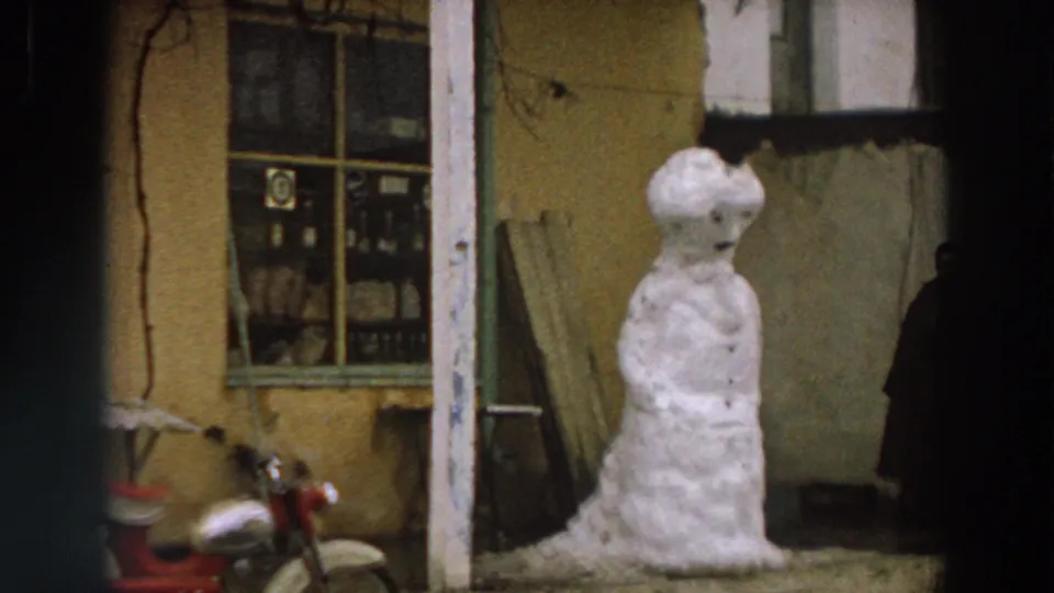 1958 Italy home-movie frame showing a snowman beside a weathered building, shop-style window, and partly visible red motorcycle.