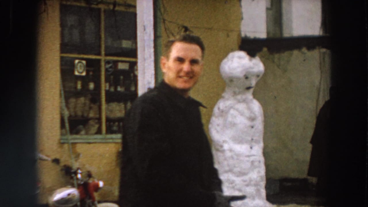 1958 Italy street frame with an adult man in a dark coat smiling toward the camera as he passes a snowman by a window.
