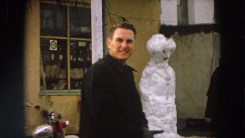 1958 Italy street frame with an adult man in a dark coat smiling toward the camera as he passes a snowman by a window.