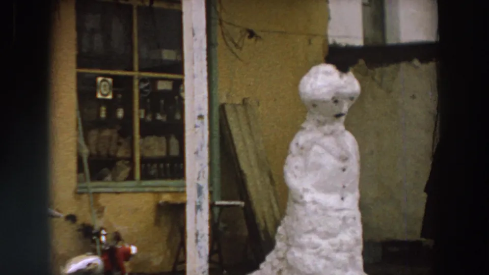 1958 Italy home-movie frame centered on a snowman near a worn wall, window display, and the edge of a red motorcycle.