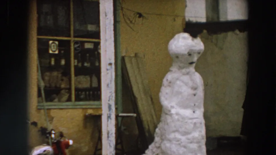 1958 Italy frame showing the snowman beside a weathered shopfront window, with a partial dark-coated figure near the edge.