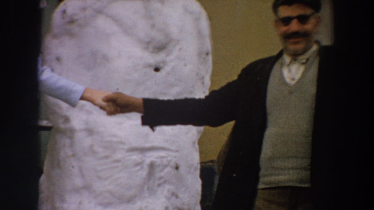 Man in flat cap and sunglasses shaking hands beside a large white sculpture.