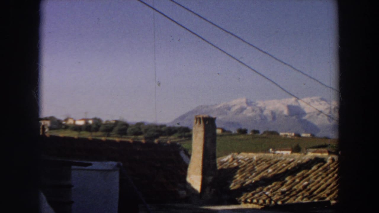 The camera looks through a window in Italy toward tiled rooftops, a stone chimney, green fields, and distant snow-capped mountains.