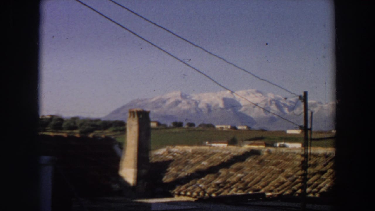 1958 Italian home-movie frame showing a tiled roof, chimney, power lines, fields, and distant snow-covered mountains.