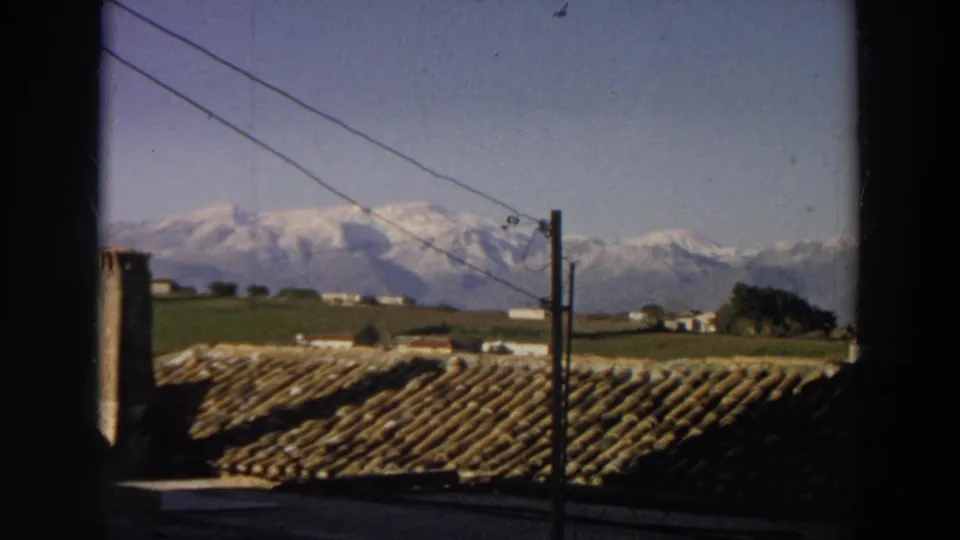 Italian 8mm home-movie still with roof tiles, a utility pole, green countryside, and a broad snow-covered mountain range.