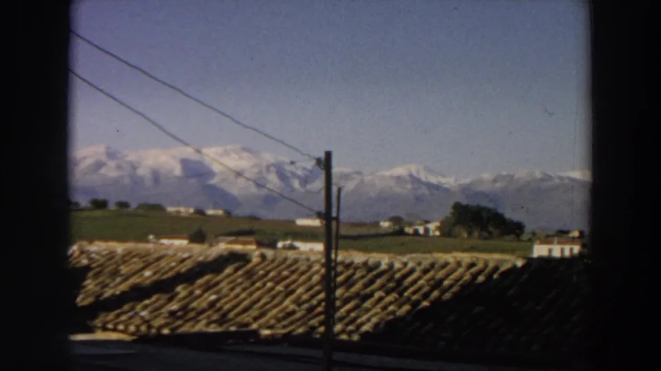 Late-frame 1958 Italy view showing terracotta roof tiles, a utility pole, fields, trees, pale buildings, and snowy mountains.