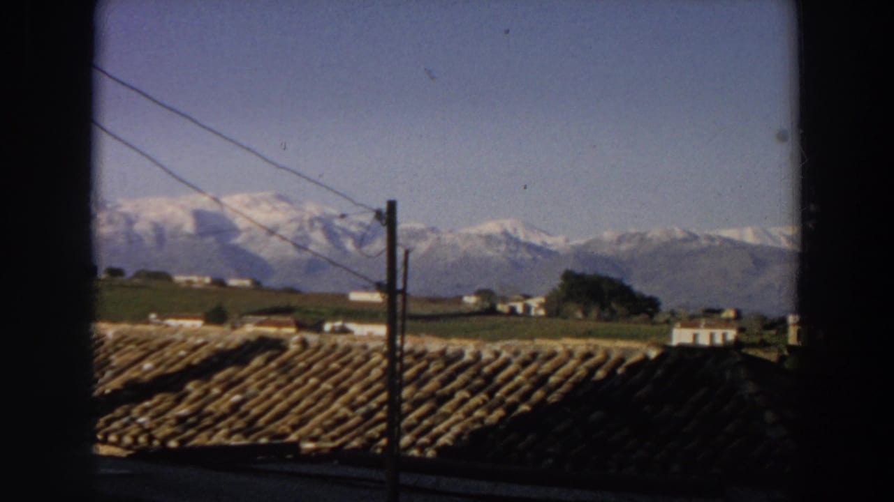 1958 Italy 8mm frame showing terracotta rooftops, a utility pole, scattered rural houses, green fields, and snow-capped mountains in the distance.