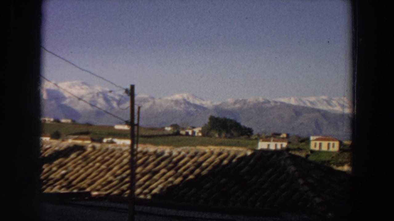 Window-framed rural landscape with rooftops, utility wires, houses, fields, and distant mountains.
