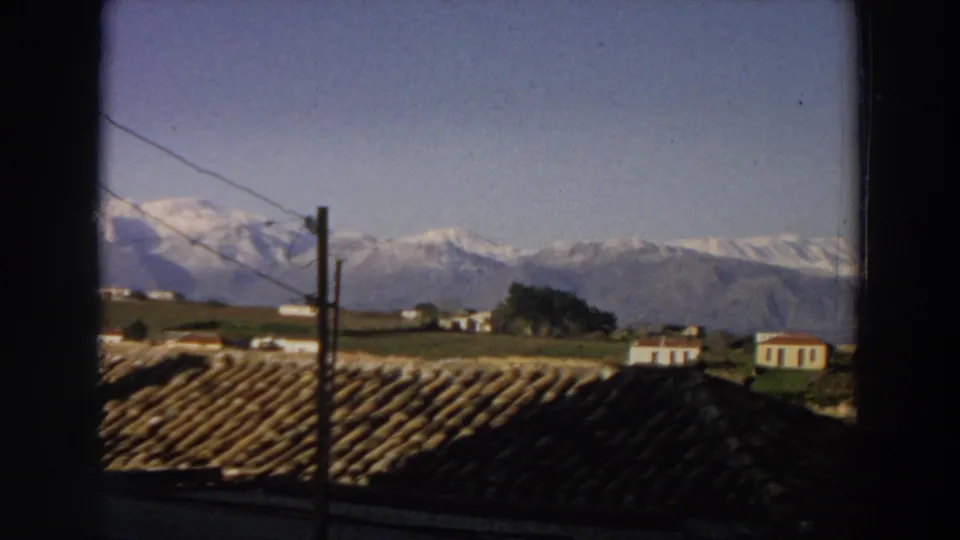 1958 Italian countryside view framed by a dark window edge, with tiled roofs, small houses, green hills, utility wires, and snowy mountains.