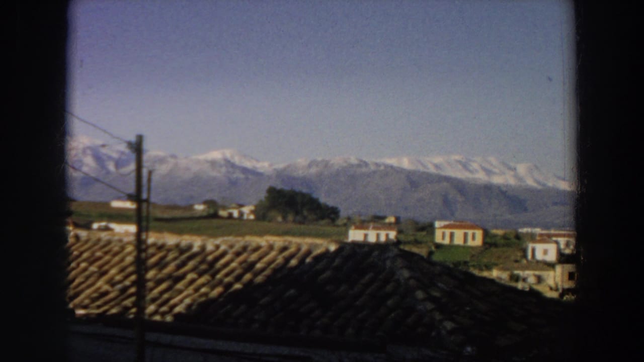 Italian 8mm home-movie frame from 1958 showing dark foreground roof tiles, rural houses, a utility pole, open fields, and a mountain range.