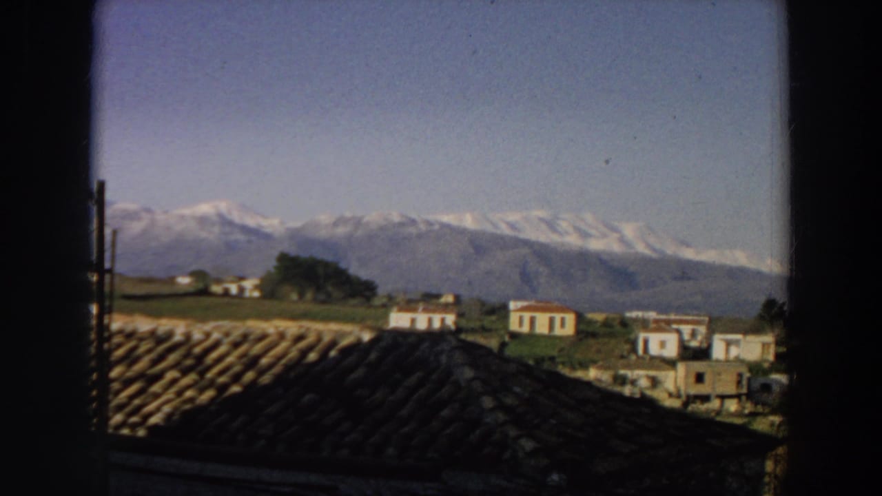 1958 Italy countryside frame with terracotta roofs, scattered white buildings, green fields, dark window edges, and distant snow-covered peaks.