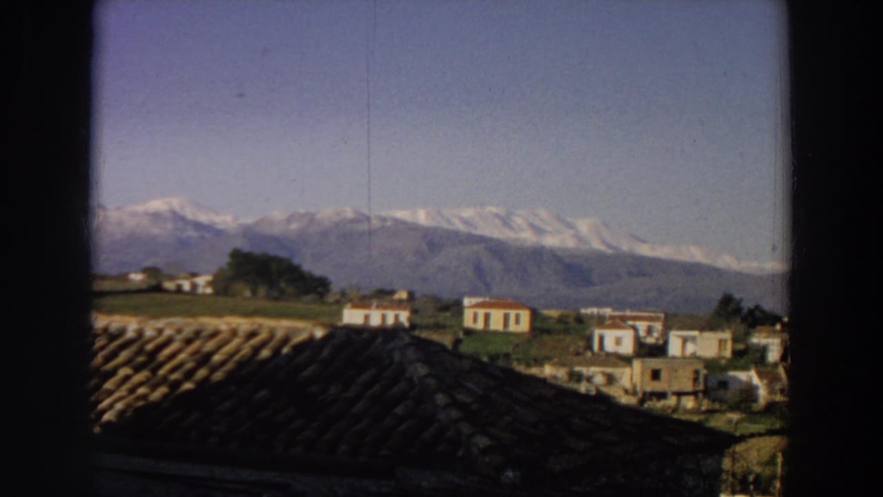 1958 Italy home-movie frame showing small white houses, a dark roof foreground, and snow-capped mountains under blue sky.