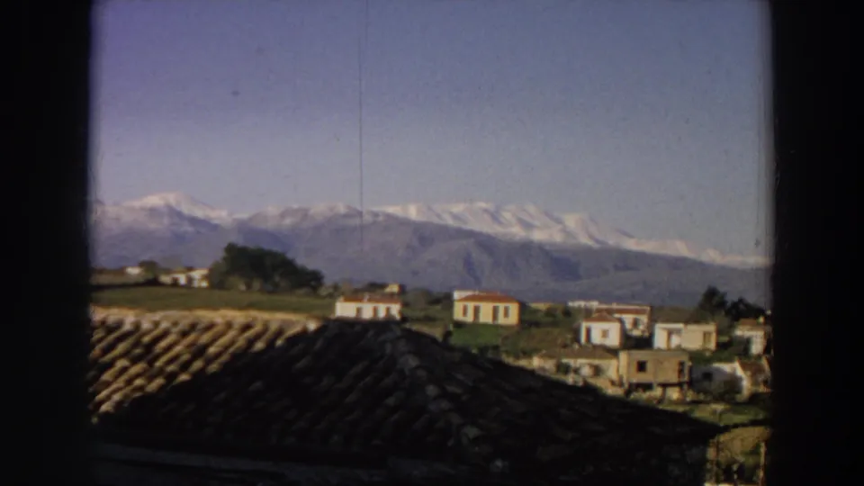1958 Italy home-movie frame showing small white houses, a dark roof foreground, and snow-capped mountains under blue sky.