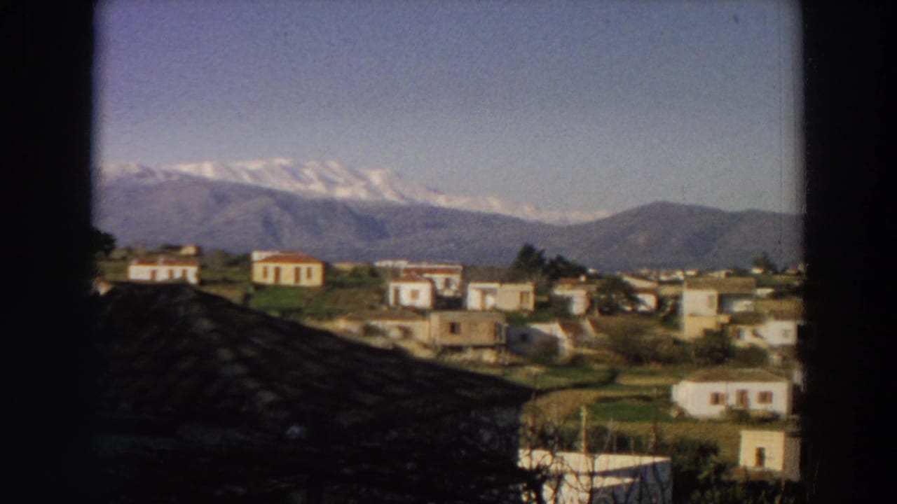1958 Italy frame showing white houses, a curving road or path, green fields, and snow-capped mountains beyond.