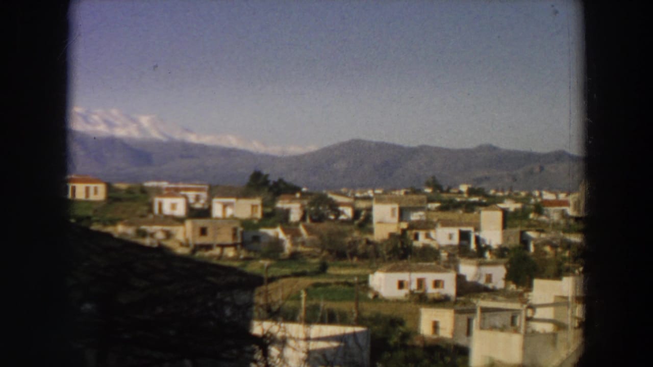 1958 Italy home-movie frame showing a hillside village of white buildings, green fields, and mountains in the distance.
