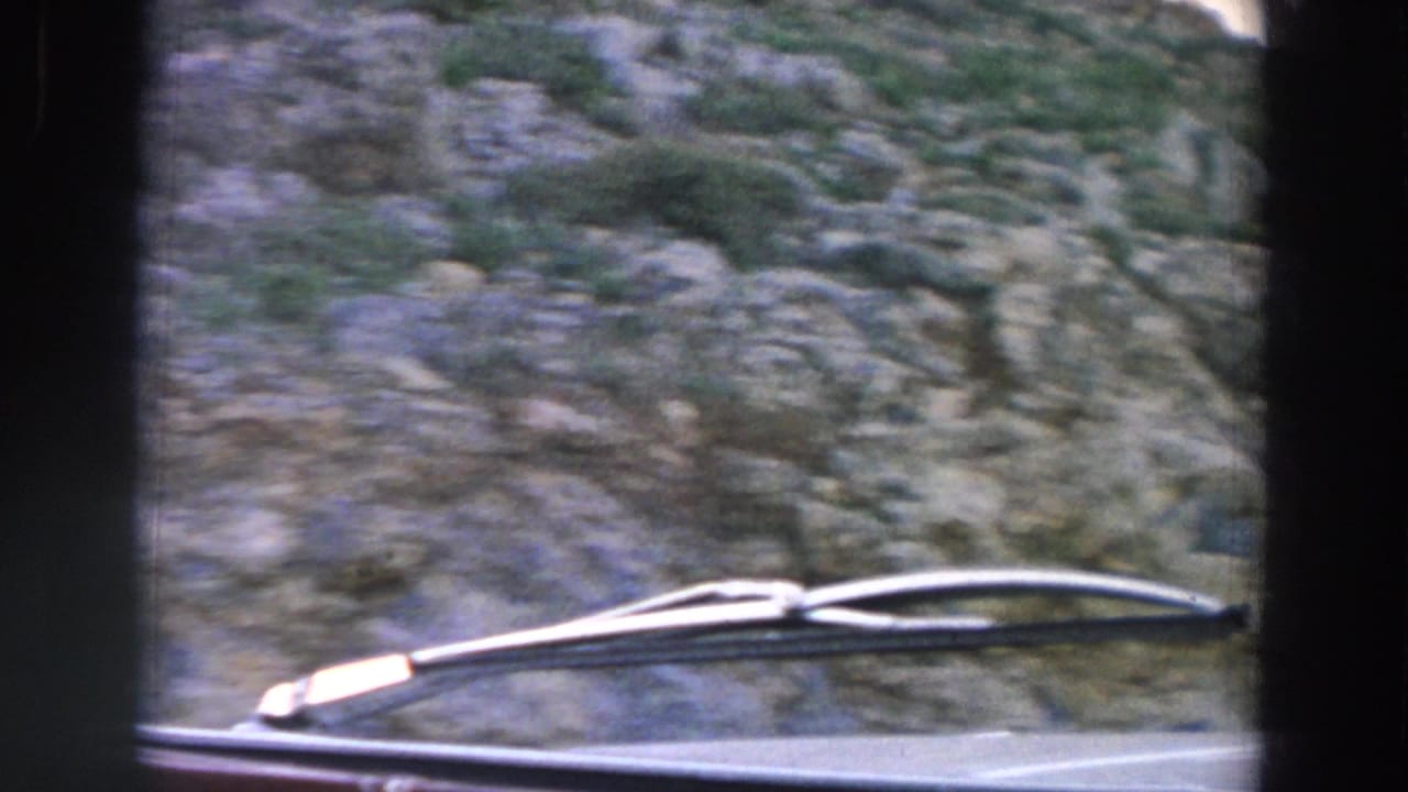 1958 Italy home-movie frame from inside a moving vehicle, showing a wiper blade against a blurred rocky roadside slope.