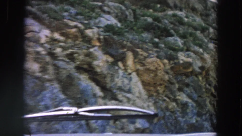 1958 8mm car-travel view in Italy, looking past windshield wipers toward a rocky roadside beside a narrow rural road.
