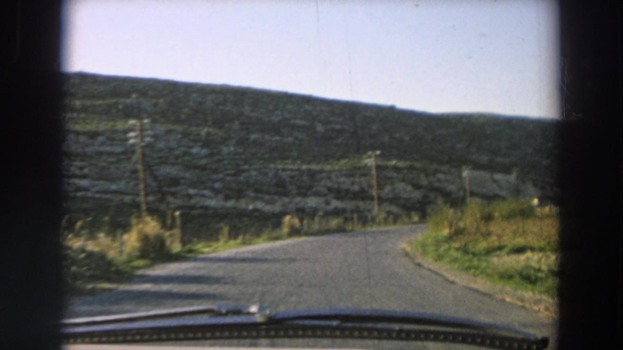 1958 Italy 8mm frame of a car windshield view along a curving rural road bordered by grass, poles, and stone hillsides.