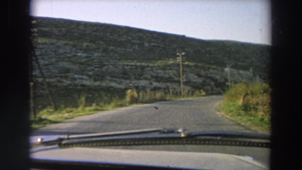 1958 Italy home-movie frame from inside a moving car, showing a quiet road curving through a stone-walled hillside landscape.