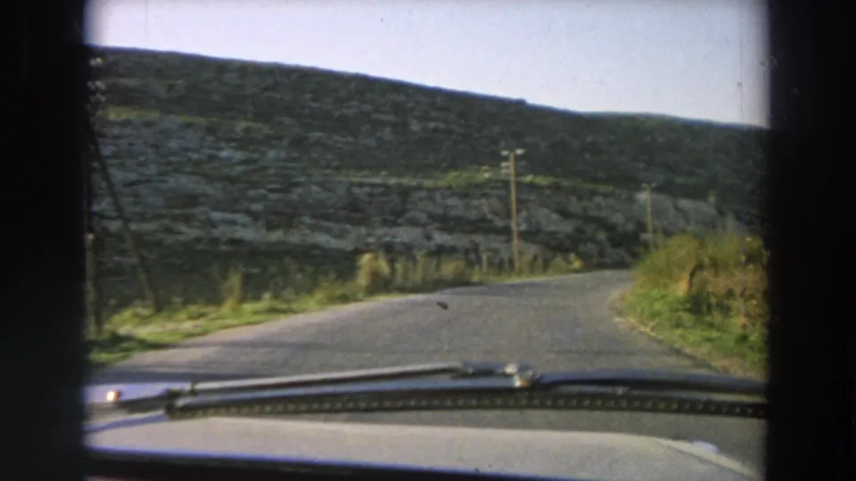 1958 Italy home-movie frame from inside a moving car, showing a quiet road curving through a stone-walled hillside landscape.