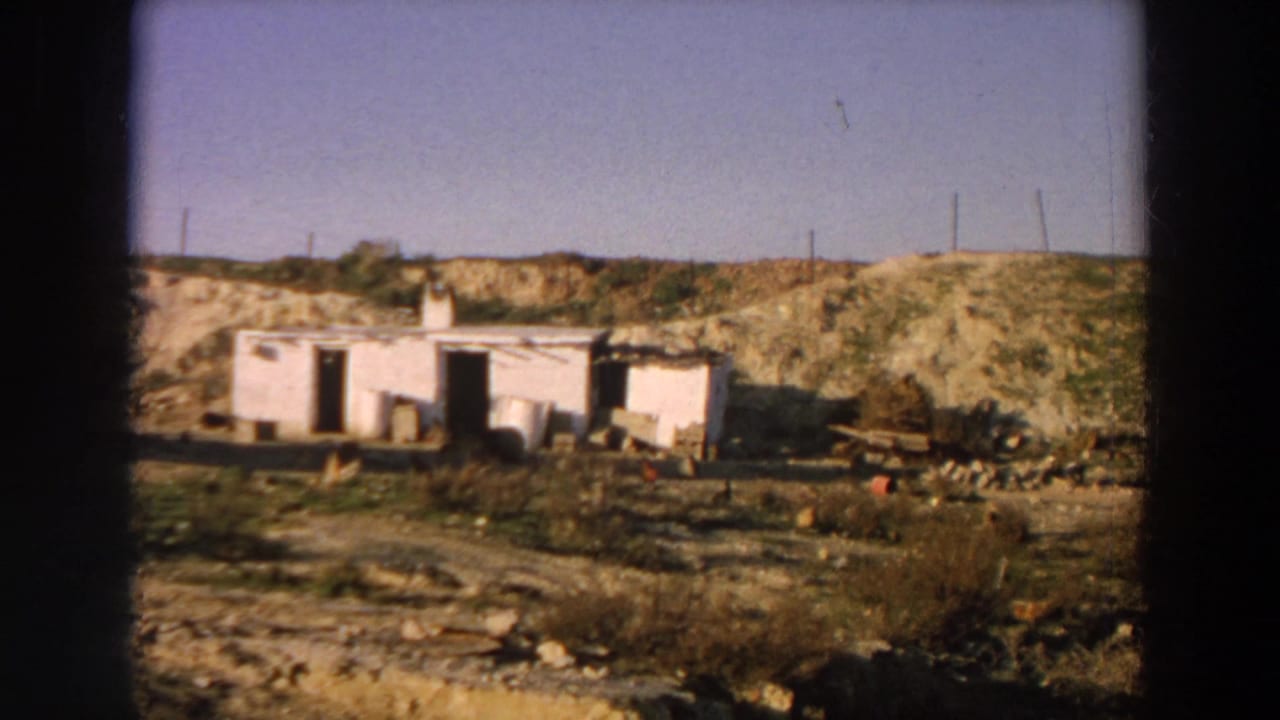 Whitewashed rural building with open doorways in a dry field, with barrels, debris, chickens, and utility poles.