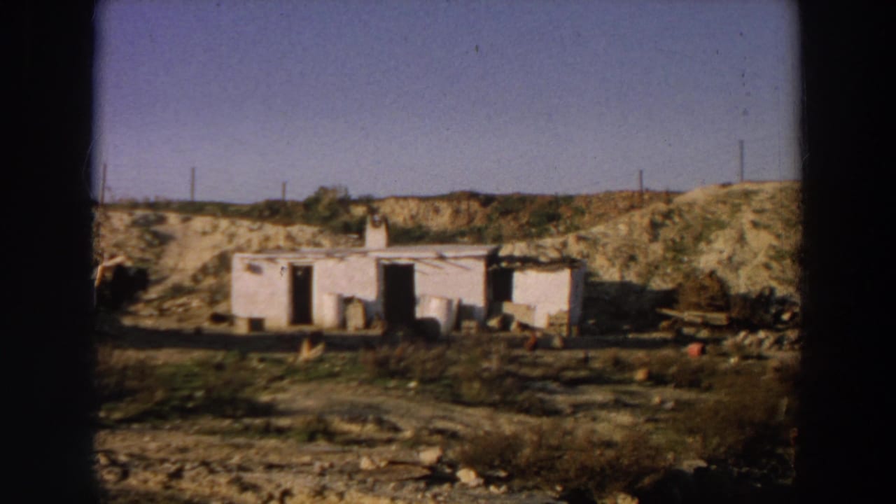 1958 Italy home-movie frame of a weathered white rural structure, open doorways, barrels, debris, sparse vegetation, and utility poles.