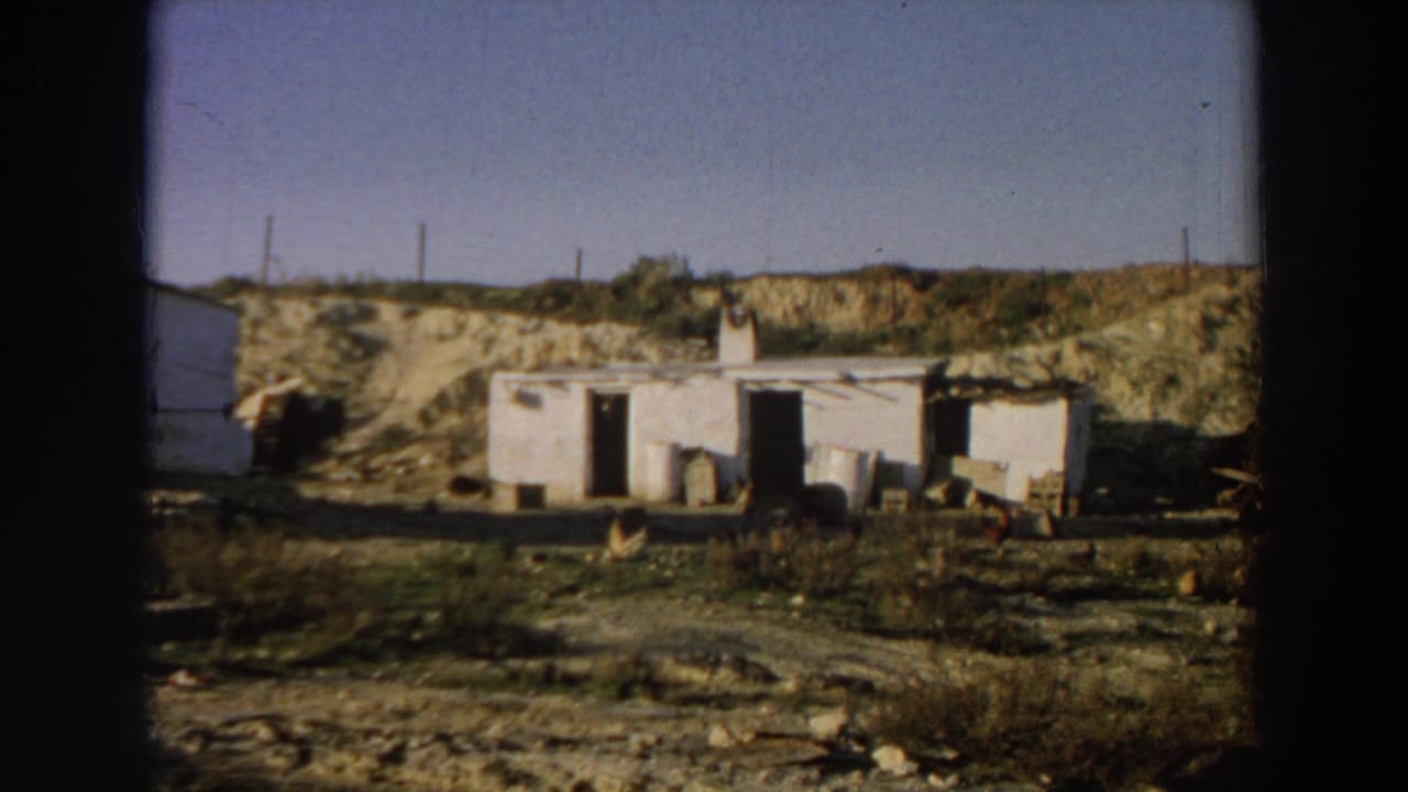 1958 Italy 8mm frame showing a whitewashed rural building in a dry landscape with open doors, scattered material, chickens, and horizon poles.