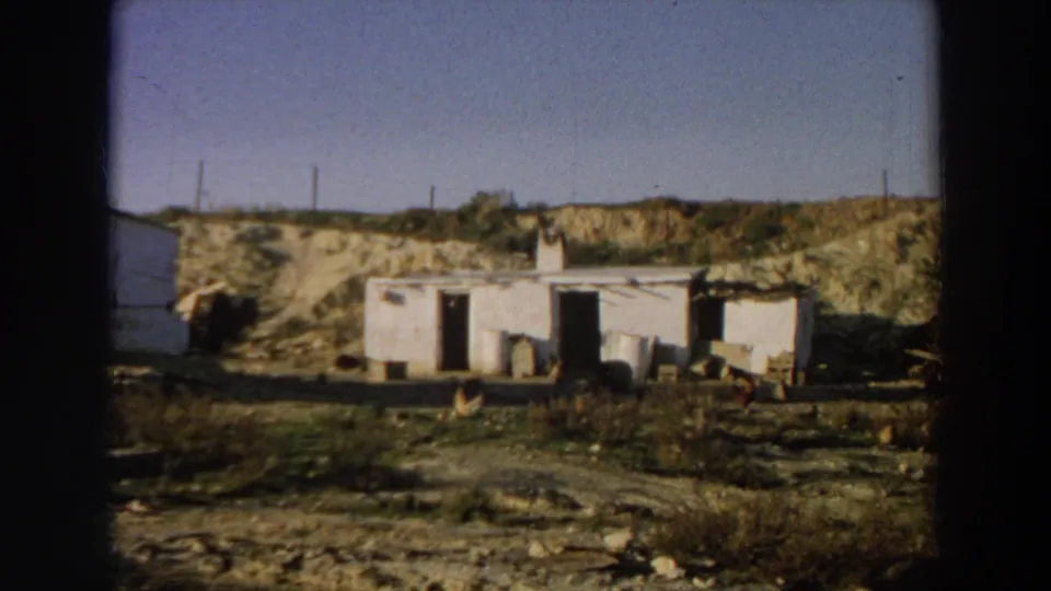 1958 Italy 8mm frame showing a whitewashed rural building in a dry landscape with open doors, scattered material, chickens, and horizon poles.