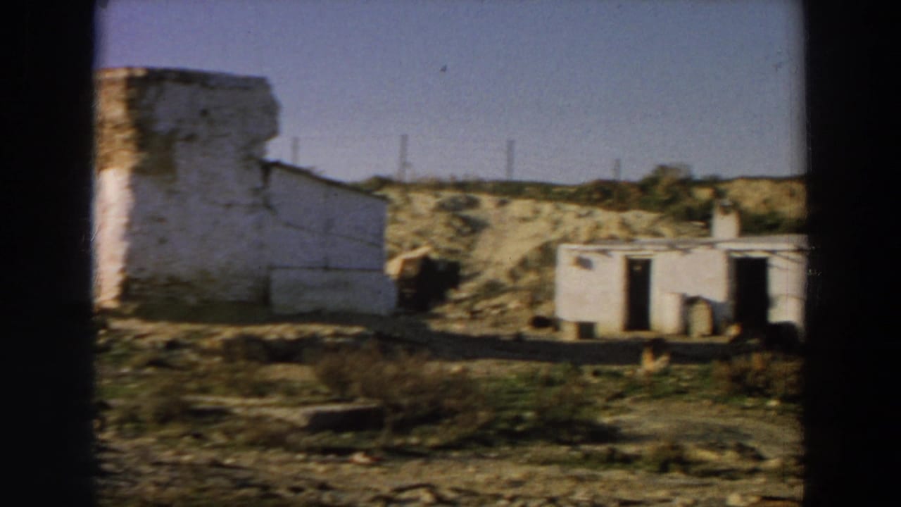 1958 Italy frame with a blurred side wall at left, a low white rural building at right, arid ground, scrub, and distant utility poles.