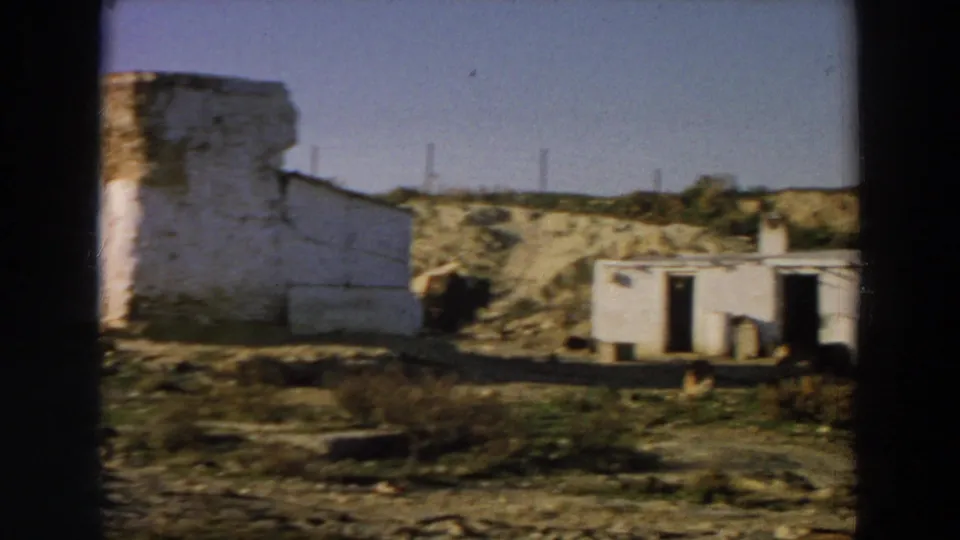 1958 Italy frame with a blurred side wall at left, a low white rural building at right, arid ground, scrub, and distant utility poles.