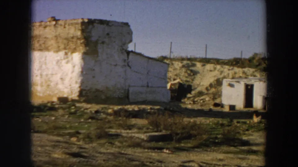 1958 Italy 8mm frame showing a weathered whitewashed ruin, dry earth, low hills, fencing, and a small shed-like structure.