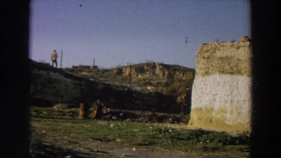 1958 Italy 8mm frame showing a distant adult figure walking along a hillside above rough ground and a whitewashed ruin.