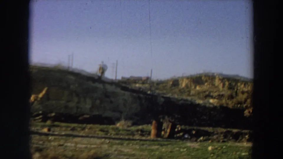 1958 Italy 8mm frame with a distant adult figure on a hillside, low ruins, dry ground, fence posts, and open sky.