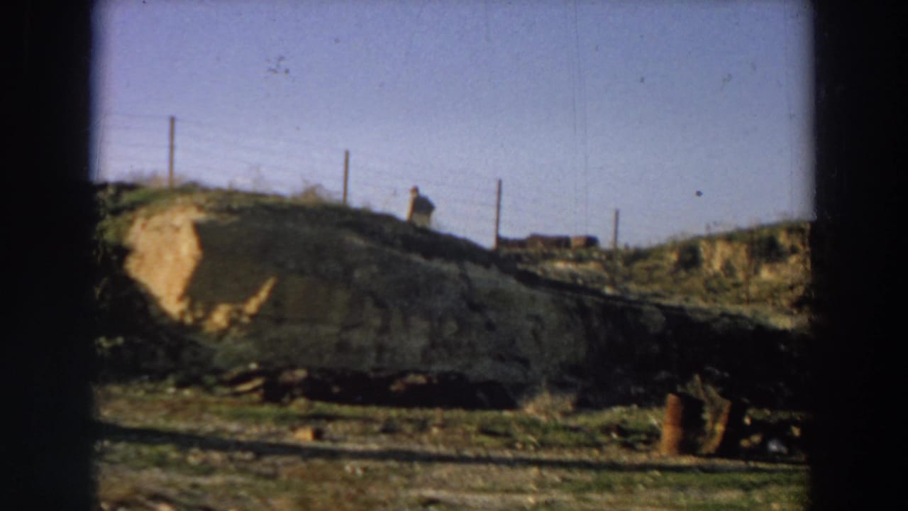 1958 Italy 8mm frame showing a rural dirt bank, fence posts, a distant walking man, and a large earthen mound by the roadside.