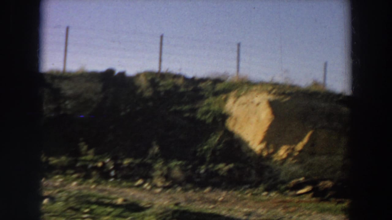 Blurred roadside view of a dirt bank, fence posts, grass, and a pale earthen mound.