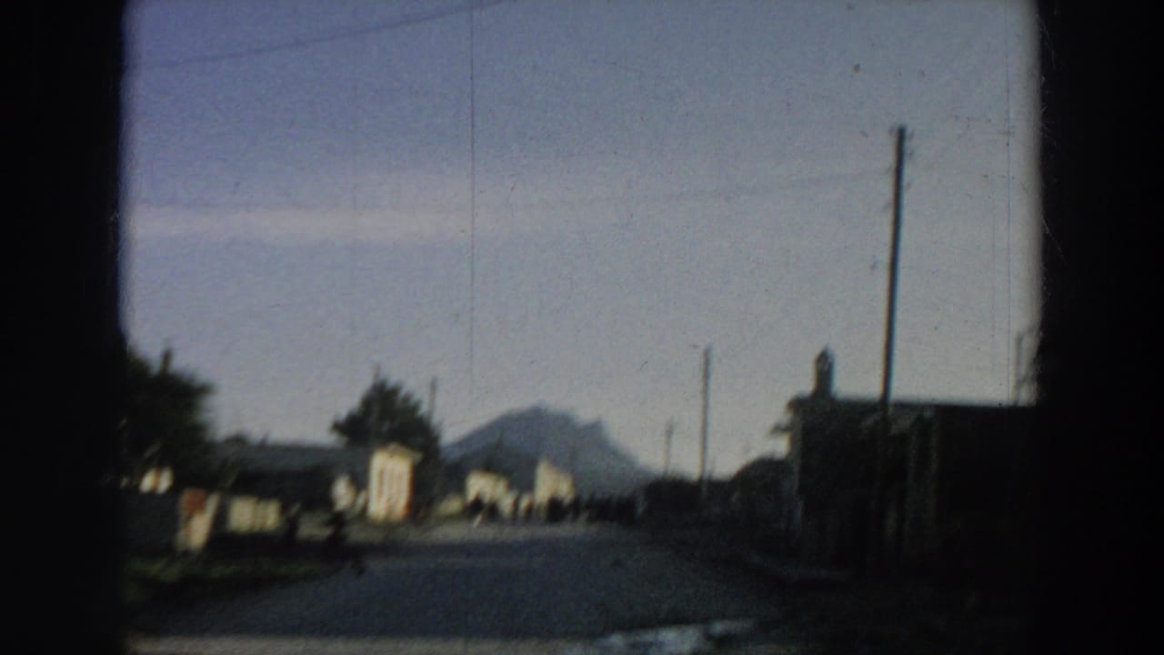 1958 Italy 8mm frame showing a quiet village road, modest roadside buildings, utility poles, and a distant mountain silhouette.