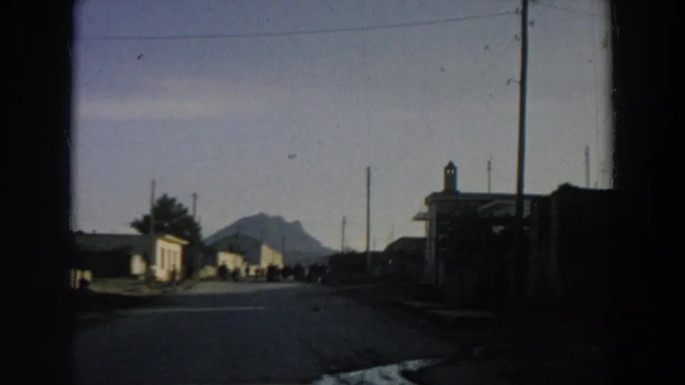 1958 Italy 8mm view from a moving car toward a small road, roadside buildings, distant mountain, and figures gathered ahead.