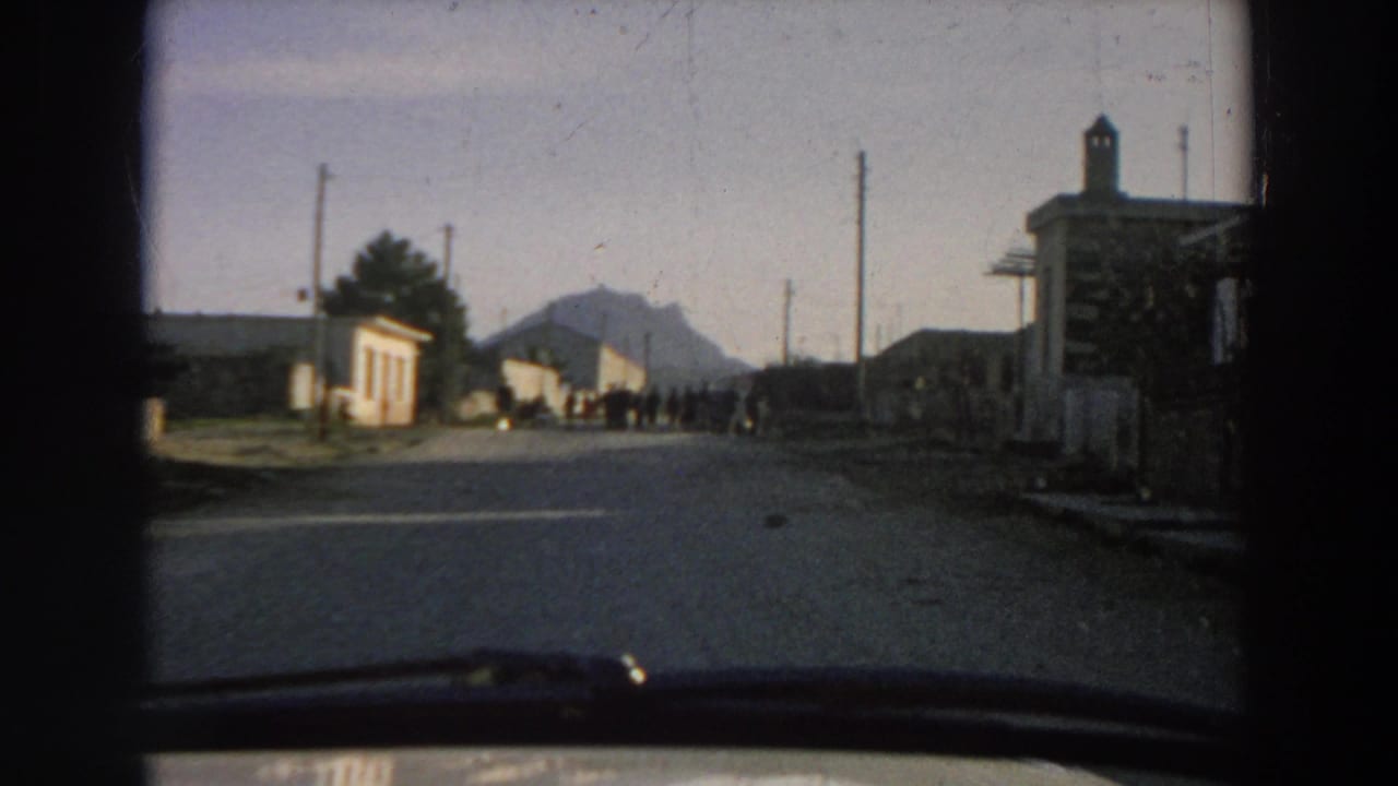 The moving-car view advances toward people standing in a road near modest buildings and a mountain backdrop in Italy.