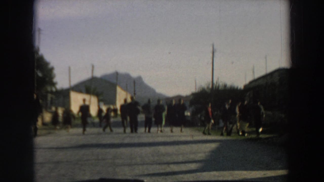 Several people stand or move across a sunlit Italian road while the car approaches, with buildings and a mountain behind them.